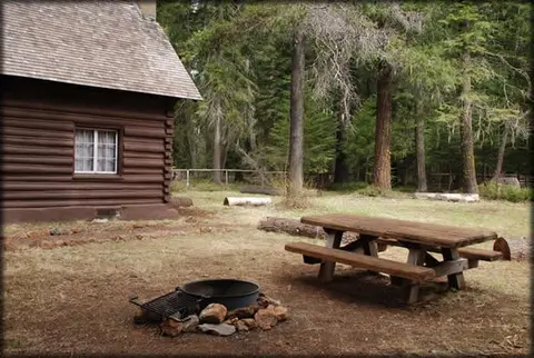A log cabin exterior with a picnic table and fire ring in a wooded area.