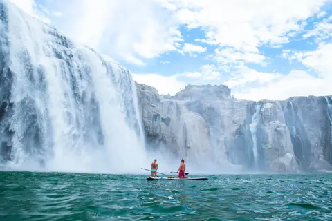 Two people paddleboarding in front of a massive waterfall.
