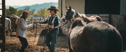 A man and a woman stand outdoors holding reins near horses by a barn.