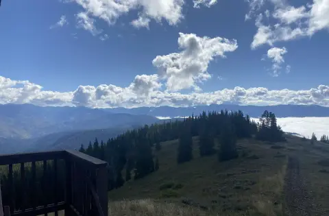 A scenic landscape view from a wooden deck overlooking mountains and forests under a cloudy blue sky.