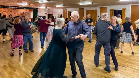 Couples dance together on a wooden floor inside a community hall.