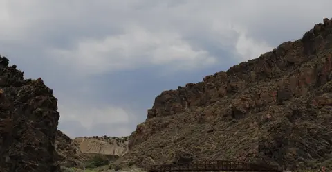 A metal bridge spans a rocky canyon under a cloudy sky.