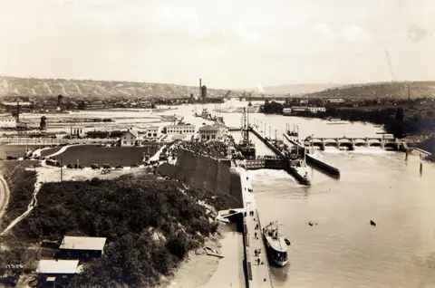 An old historic photograph shows a canal with locks, boats, and a crowd of people.
