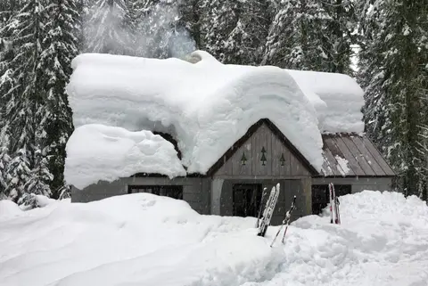 A cabin covered in deep snow is surrounded by evergreen trees.
