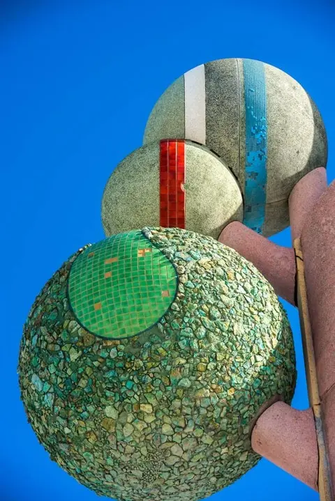 A close-up view of a sculpture with three spherical elements decorated with stones and tiles against a blue sky.