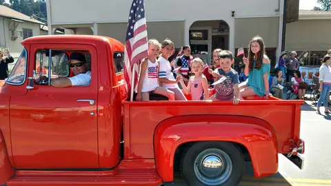 Children ride in the back of a red pickup truck holding American flags.