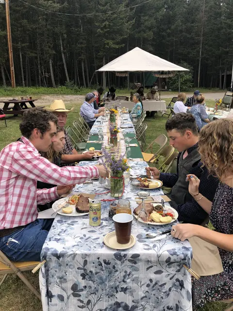 People seated at a long outdoor dining table in a forest setting.
