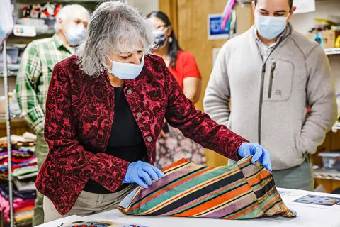 People in masks examine a striped fabric item indoors.