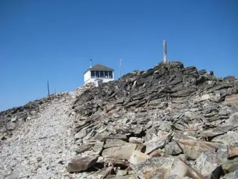 A small cabin sits on a rocky mountain summit under a clear blue sky.