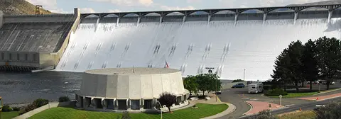 A large dam with water cascading over its spillway behind a round building and parking area.