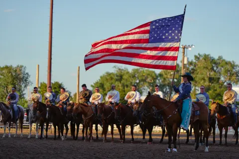 A person on horseback carries a large American flag while a line of people on horseback hold their hats in front of them.