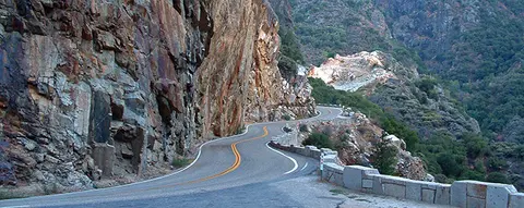 A winding mountain road passes next to rocky cliffs and a stone wall.
