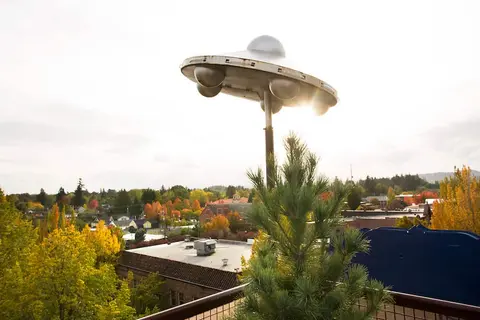 An outdoor sculpture of a flying saucer stands above a town with colorful autumn trees.