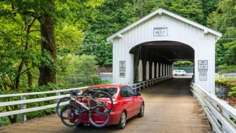 A red car with bicycles mounted on the back is parked in front of a white covered bridge.