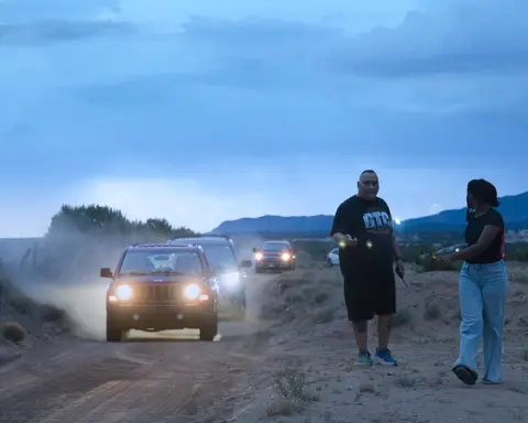 Two people stand on the side of a dusty road as several vehicles approach with headlights on at dusk.