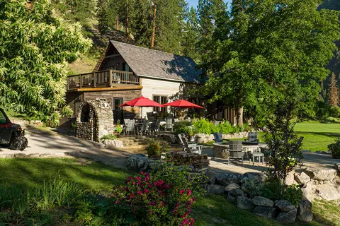 A rustic wooden building with an outdoor patio, red umbrellas, and a fire pit area surrounded by trees and mountains.