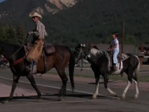 A cowboy and a child ride horses down a street with mountains in the background.