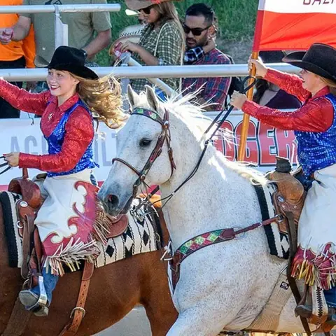 Two women in western wear ride horses at an event.