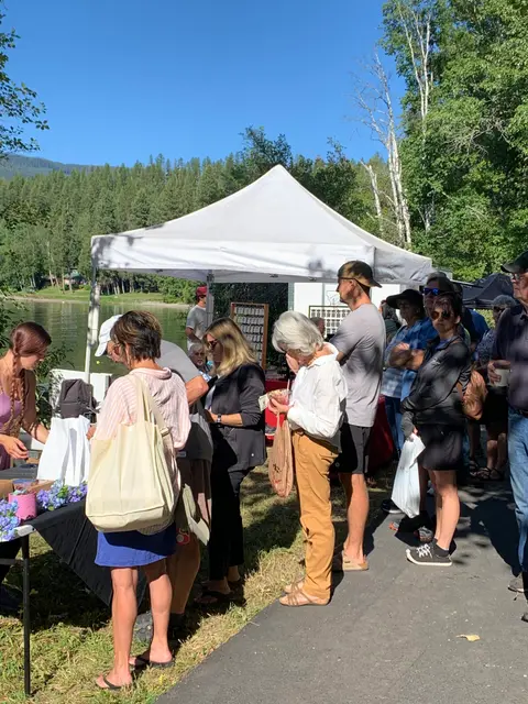 People wait in line at an outdoor market tent near a lake.