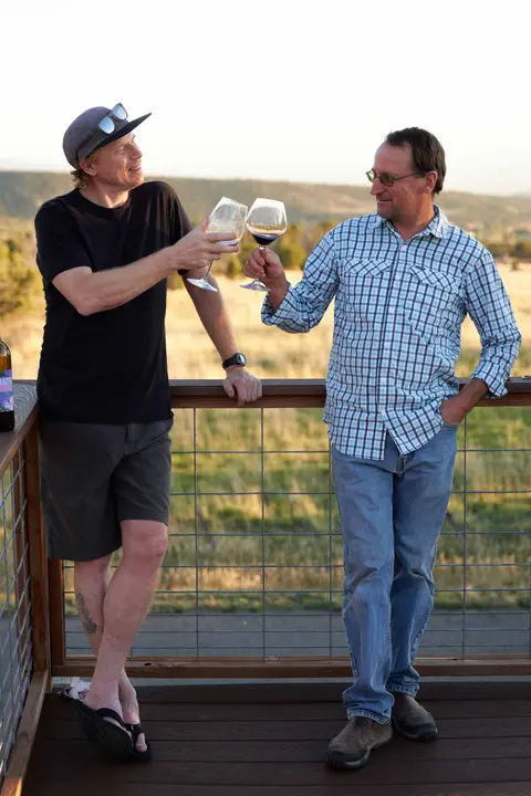 Two men toast each other with wine glasses on an outdoor deck overlooking a landscape.