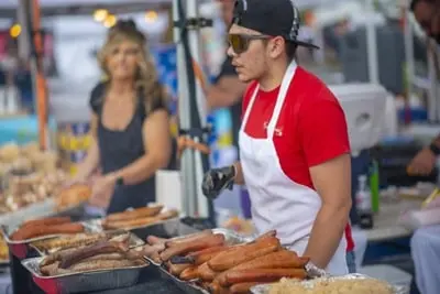 A vendor wearing an apron and cap stands behind trays of grilled sausages at an outdoor event.