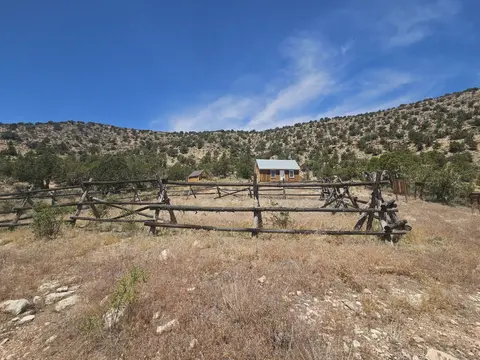A small wooden cabin stands behind a rustic wooden fence in a dry, mountainous landscape.