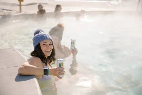 Two women smile while relaxing in a steamy outdoor hot spring pool holding drinks.