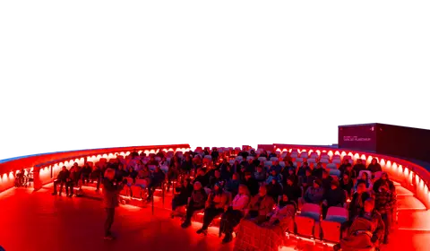 People seated inside a theater listening to a speaker under a domed ceiling.