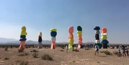 People walking around large brightly colored stacked boulders in a desert landscape.