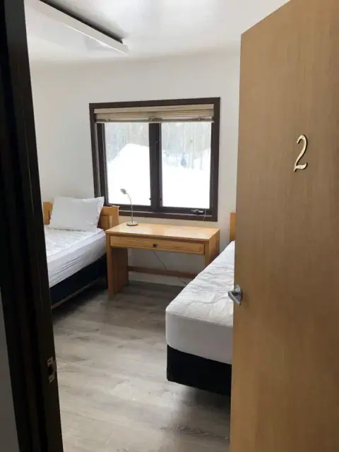 A view into a small bedroom with two beds and a desk beneath a window.