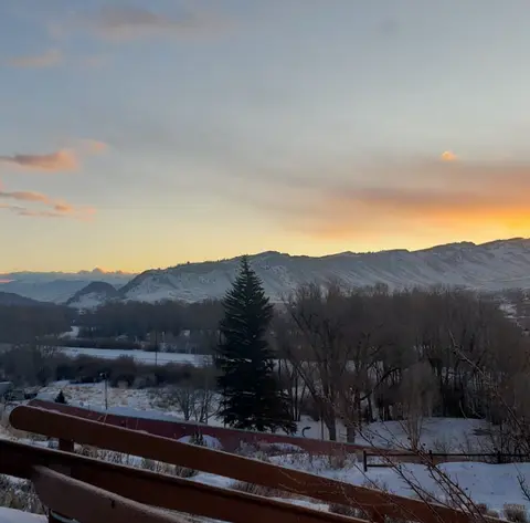 A snowy landscape with mountains and a sunset sky viewed behind trees and a wooden railing.