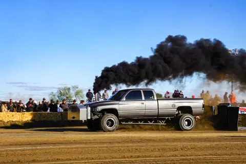 A pickup truck emits thick black smoke while pulling a sled at a truck pull event.