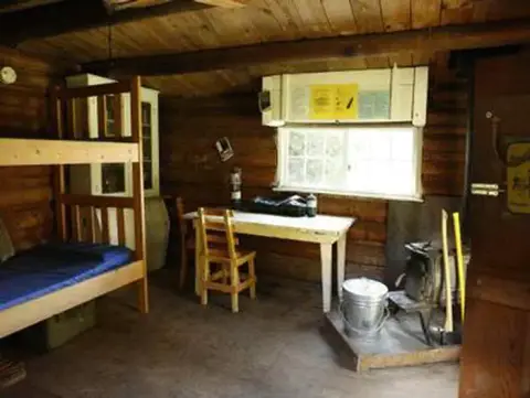 The interior of a rustic wooden cabin with a bunk bed, table, and wood stove.