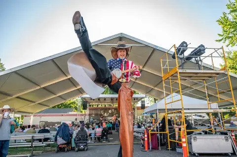 A performer on stilts dressed in a cowboy hat and American flag shirt plays a banjo at an outdoor event.