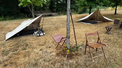 A campsite features canvas lean-to tents and wooden chairs set up on dry grass.