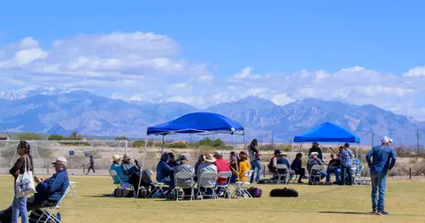 A group of people gather under pop-up tents on a grassy field with mountains in the background.