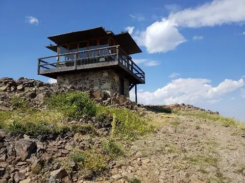 A stone and wood fire lookout tower stands on a rocky mountain summit under a blue sky.