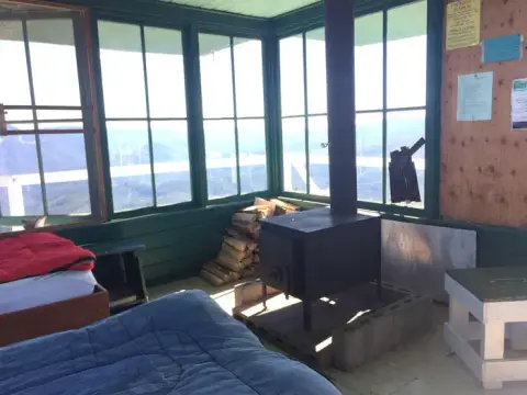 The interior of a fire lookout cabin with a wood stove and large windows.