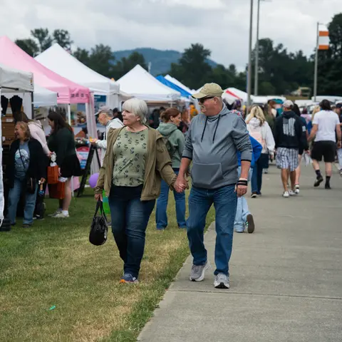 A couple walks hand-in-hand along a pathway at an outdoor fair.