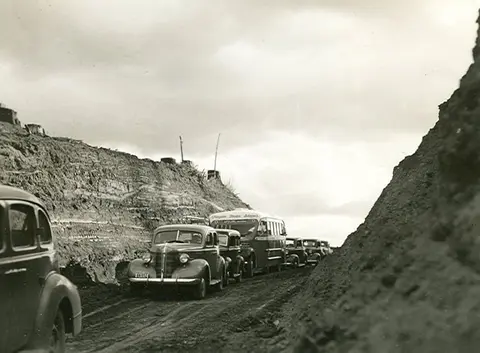 A line of vintage cars and a bus driving through a dirt cut in the earth.