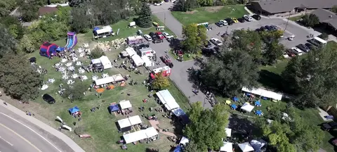 An aerial view shows an outdoor community event with tents, tables, and parked cars.
