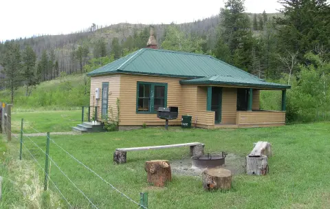 A single-story wooden building with a green roof stands in a grassy clearing with a fire pit in the foreground.
