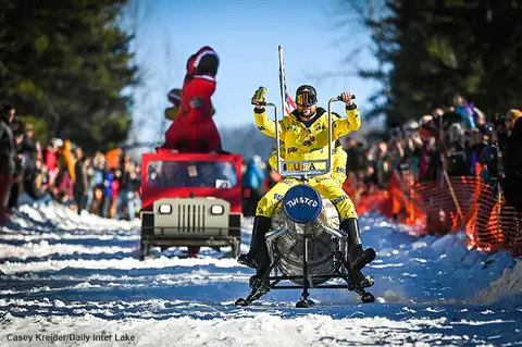 People ride a homemade sled down a snowy hill during an event.