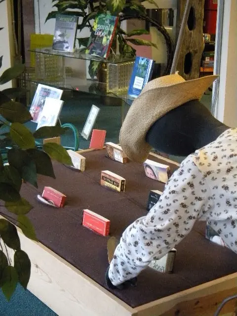 A mannequin in a cowboy hat and floral shirt reaches toward books displayed on a low brown table indoors.