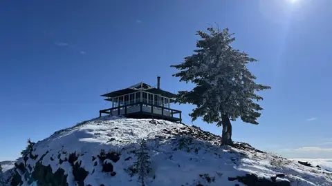 A fire lookout tower sits atop a snowy peak next to a tree under a blue sky.
